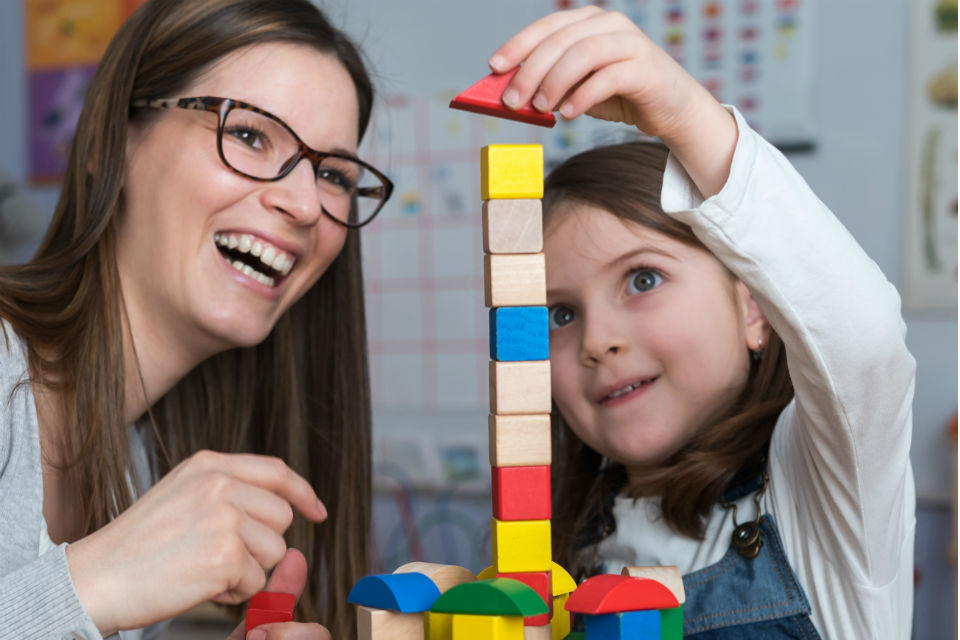 Female early years carer watching young girl build tower with toy bricks